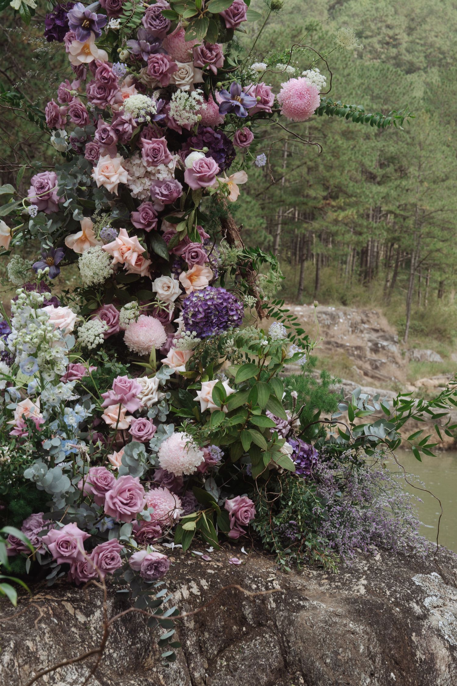 An elaborate floral arrangement of purple roses, hydrangeas, and mixed blooms cascades over a rocky ledge in a forest.