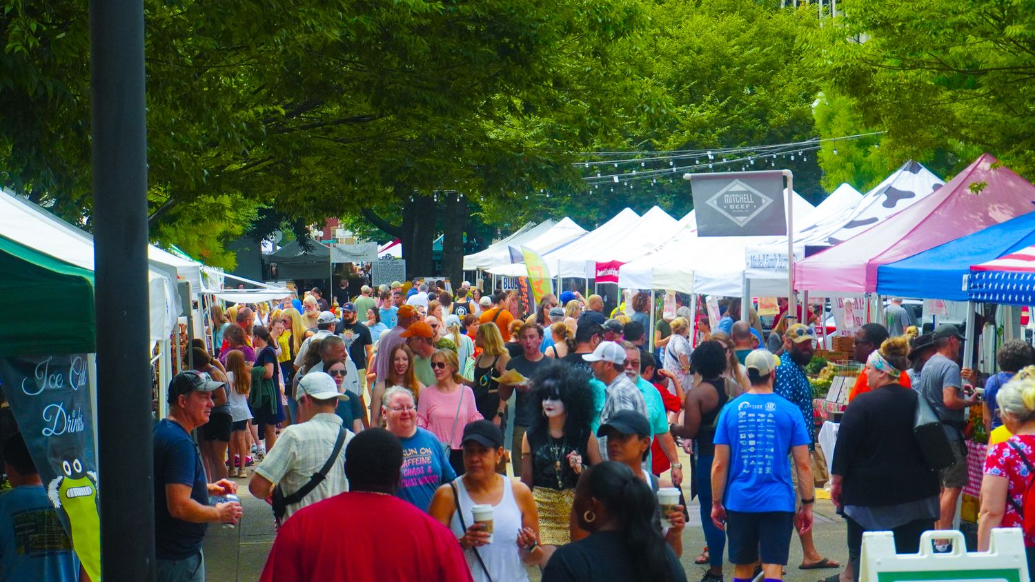 A crowded outdoor festival marketplace with white tents and vendors displaying their goods in a park-like setting.