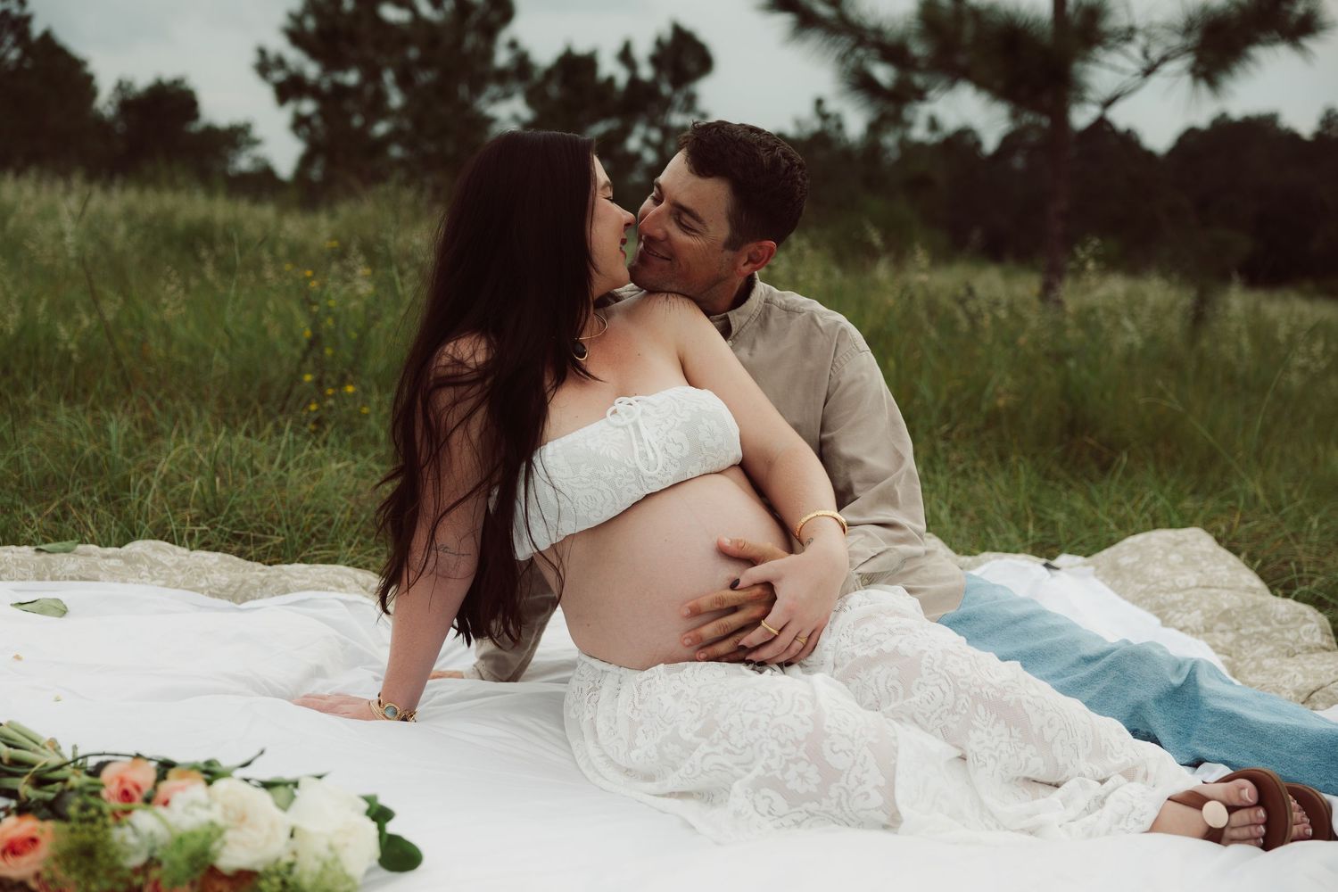 Couple snuggling together during a romantic outdoor photoshoot on a blanket with flowing white fabric and pine trees.