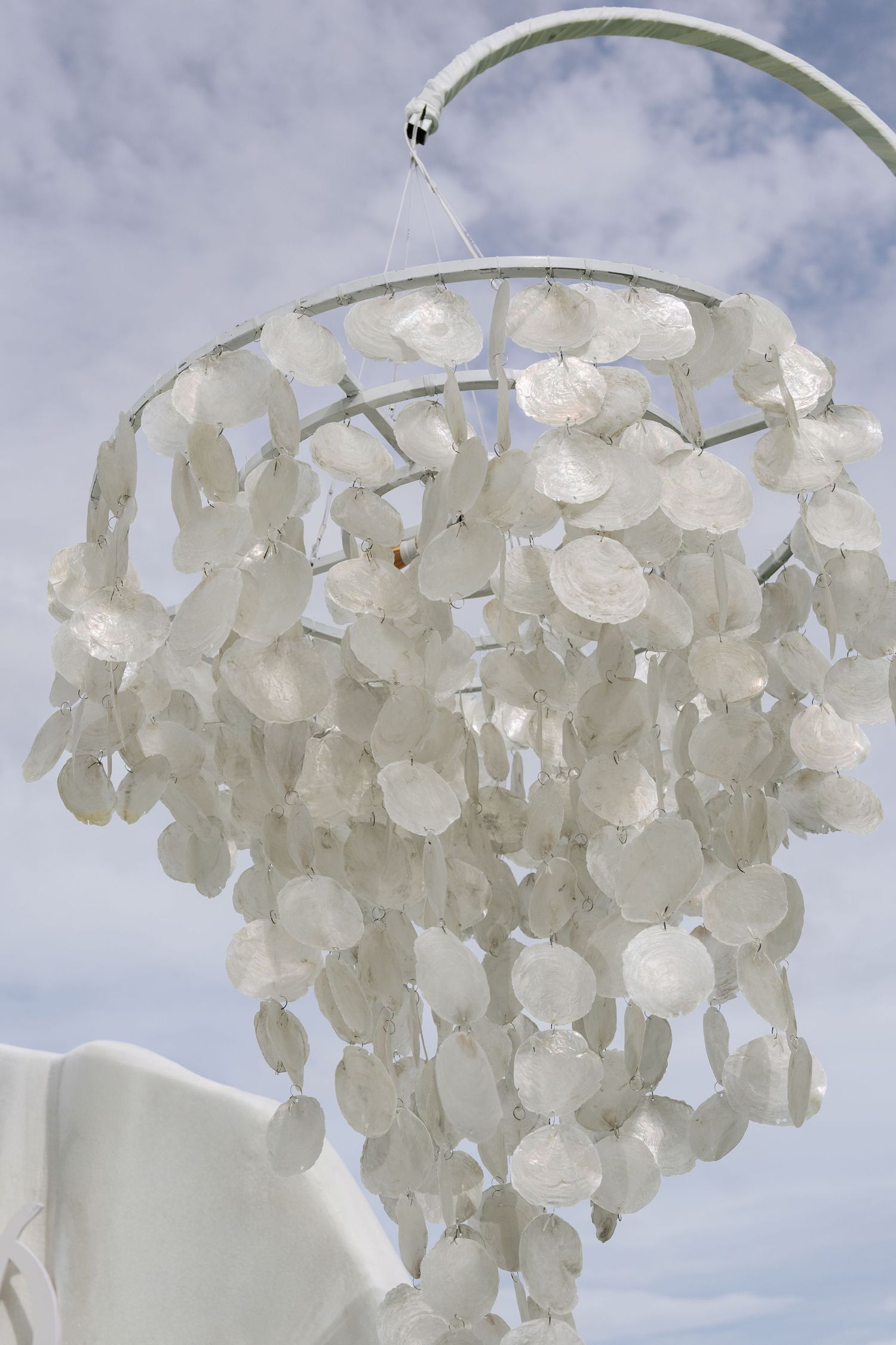 Elegant white capiz shell chandelier hanging against blue sky at an outdoor wedding ceremony.