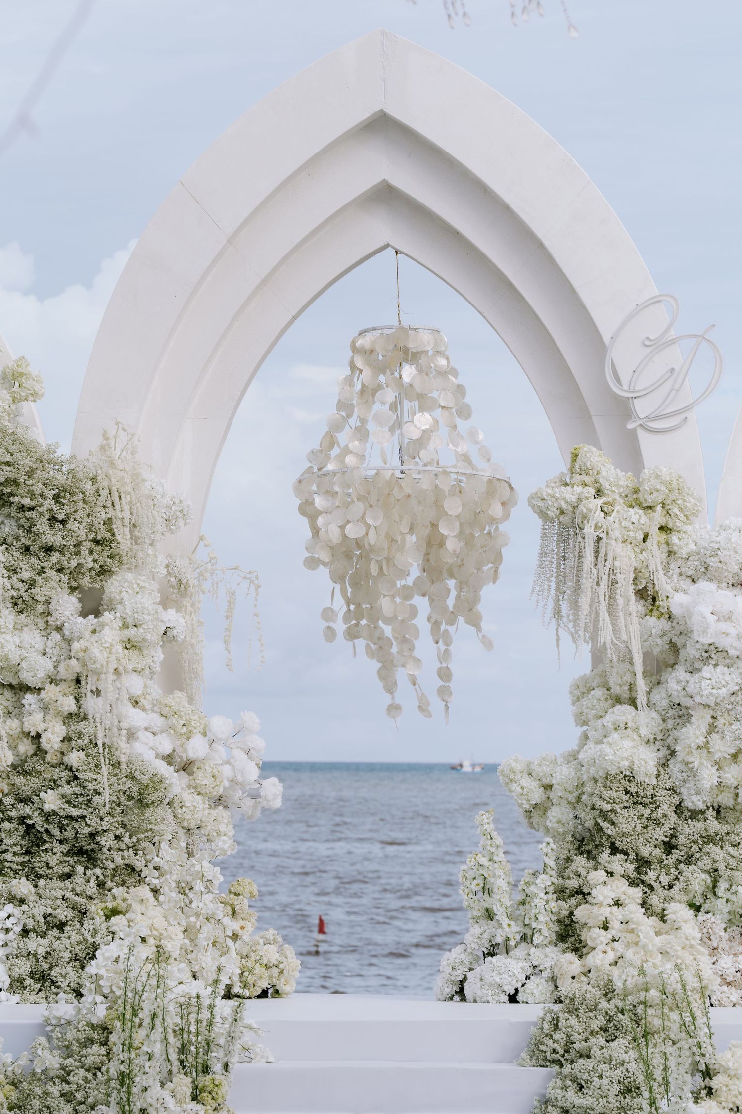 Elegant white wedding arch adorned with lush white flowers and pearl chandelier overlooking the ocean.
