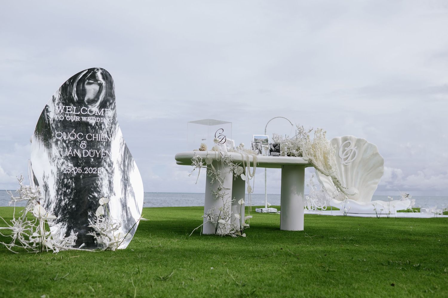 Outdoor coastal wedding setup with mirrored welcome sign, white arch, and decorated table on green lawn.