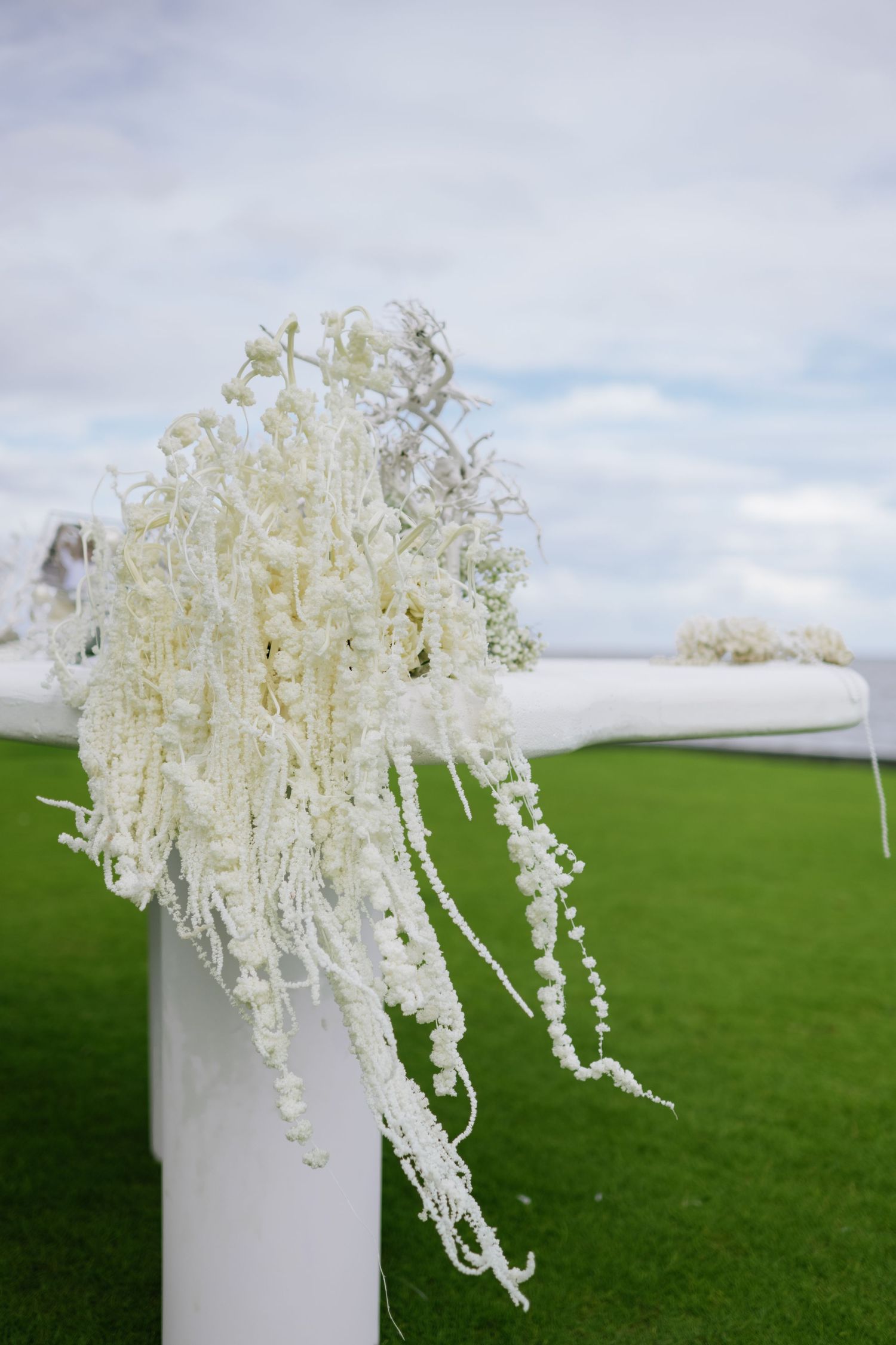 White cascading floral arrangement on a white pillar at an outdoor wedding ceremony with green lawn backdrop.