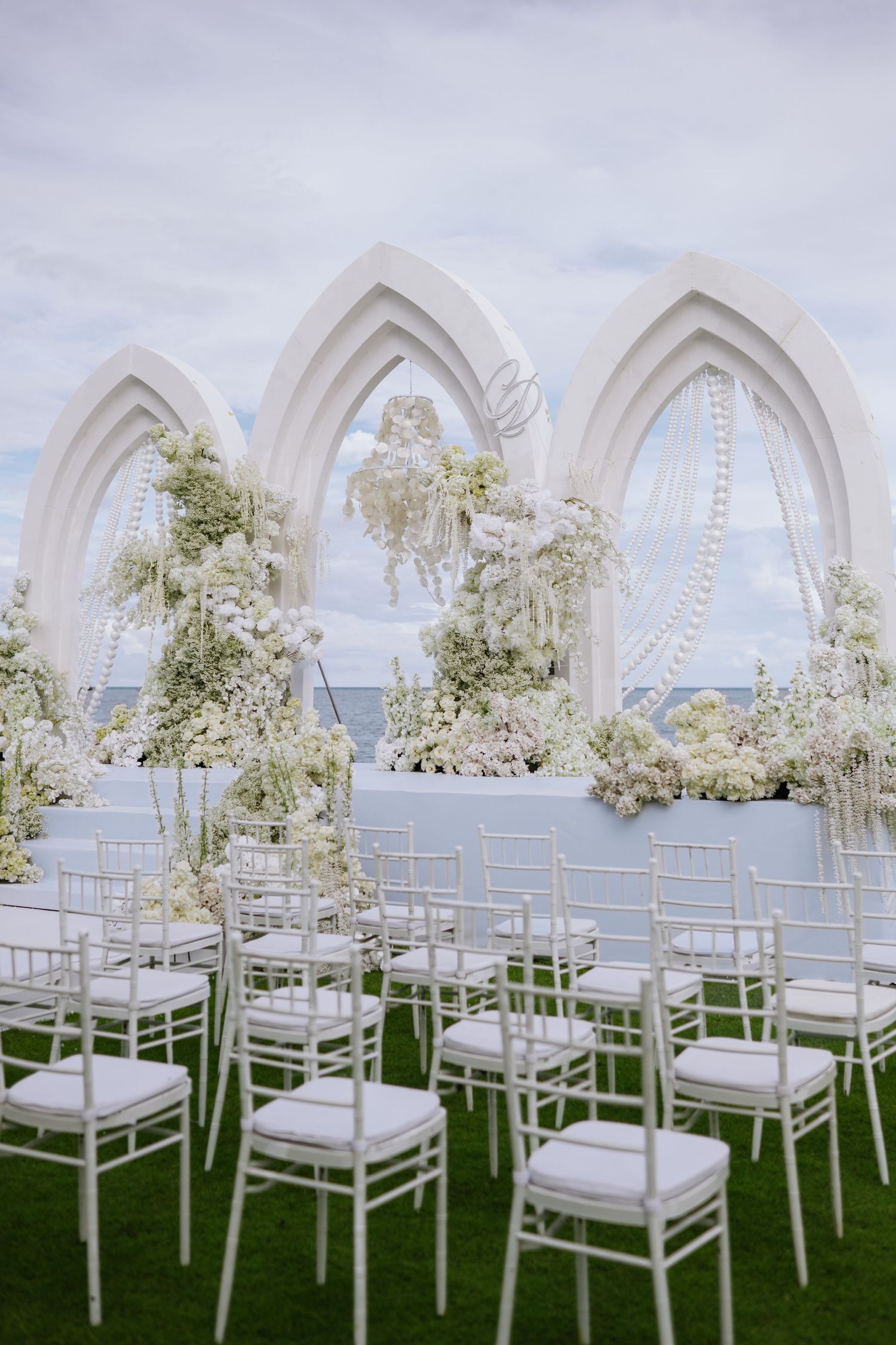 Elegant all-white outdoor wedding ceremony setup with Gothic arch backdrop, chiavari chairs, and ocean view.