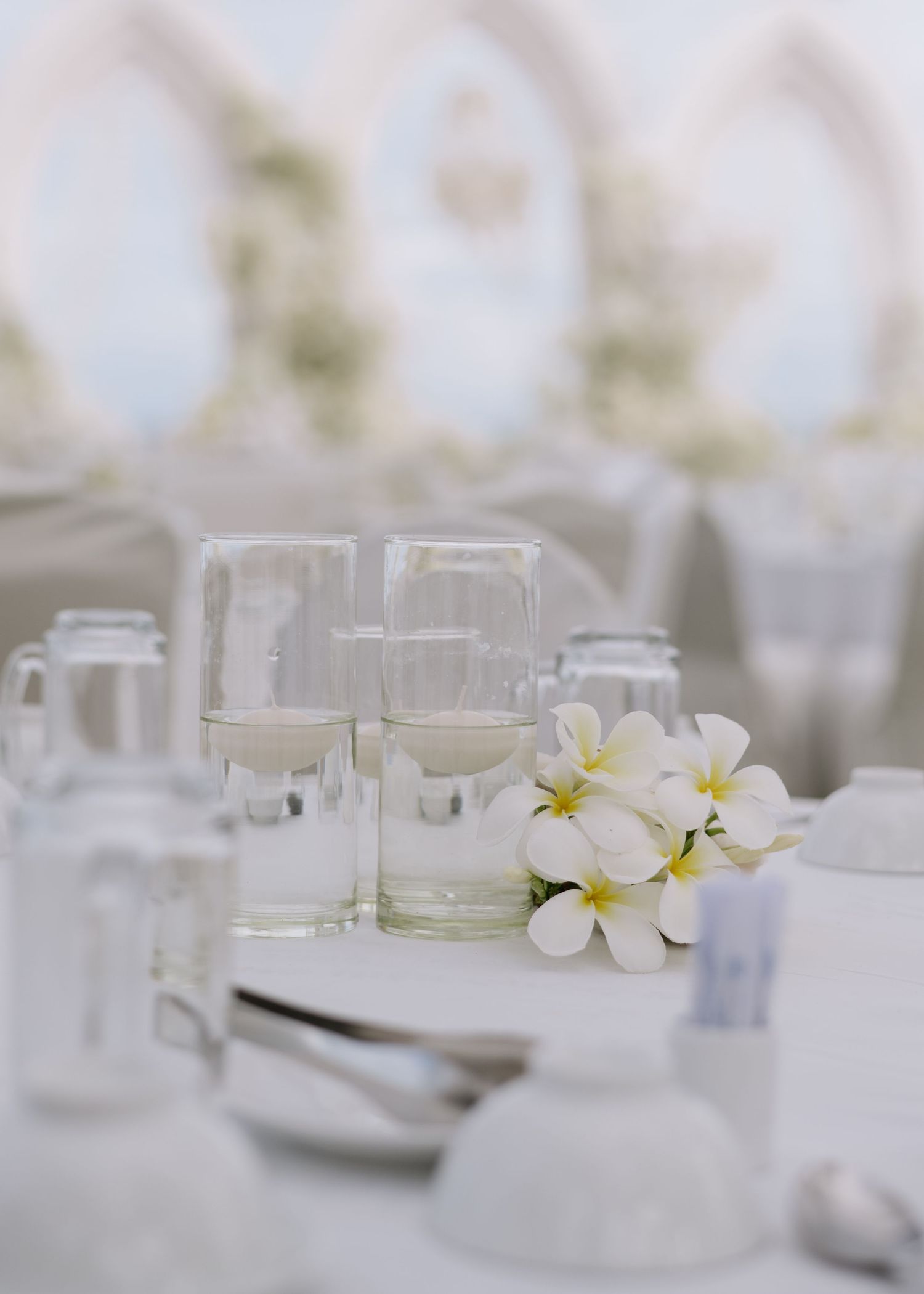 White wedding reception table setting with plumeria flowers, glassware, and soft bokeh background.