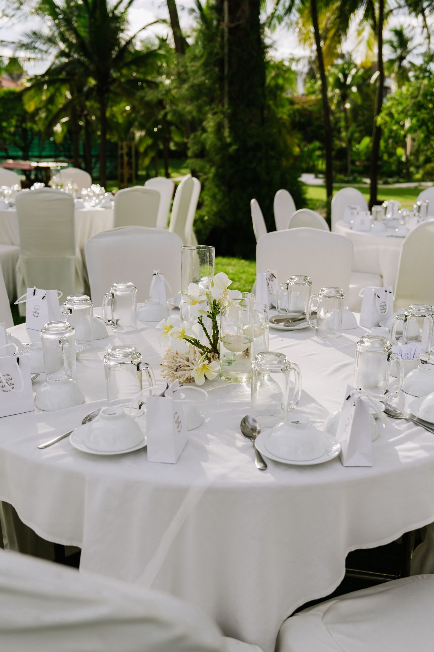 Elegant outdoor wedding reception with round white tables, white chairs, floral centerpieces, and tropical garden backdrop.