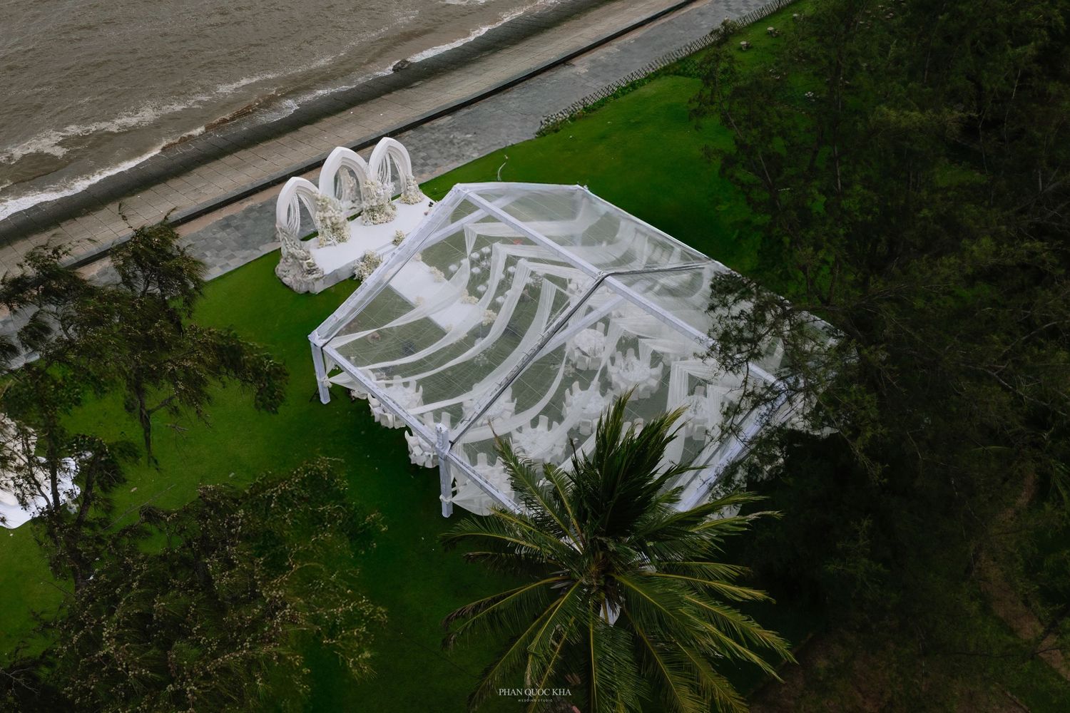 Aerial view of white wedding tent and ceremony setup on green lawn beside ocean shoreline.
