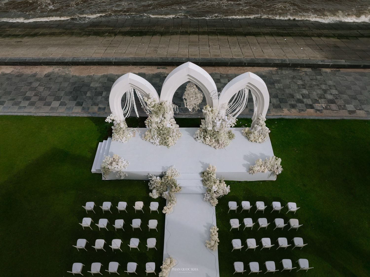 Aerial view of all-white outdoor wedding ceremony with Gothic arches, aisle, and rows of chairs.