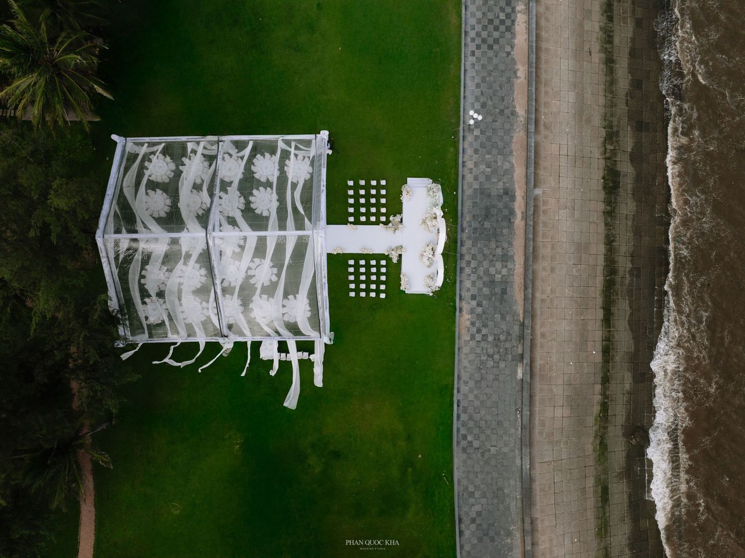 Aerial top-down view of white wedding reception tent structure on green lawn near beach.