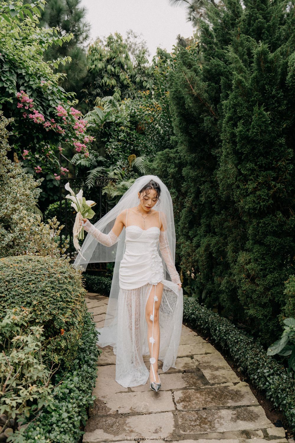 Bride in a short white wedding dress and sheer overlay strolling along a lush green garden path.
