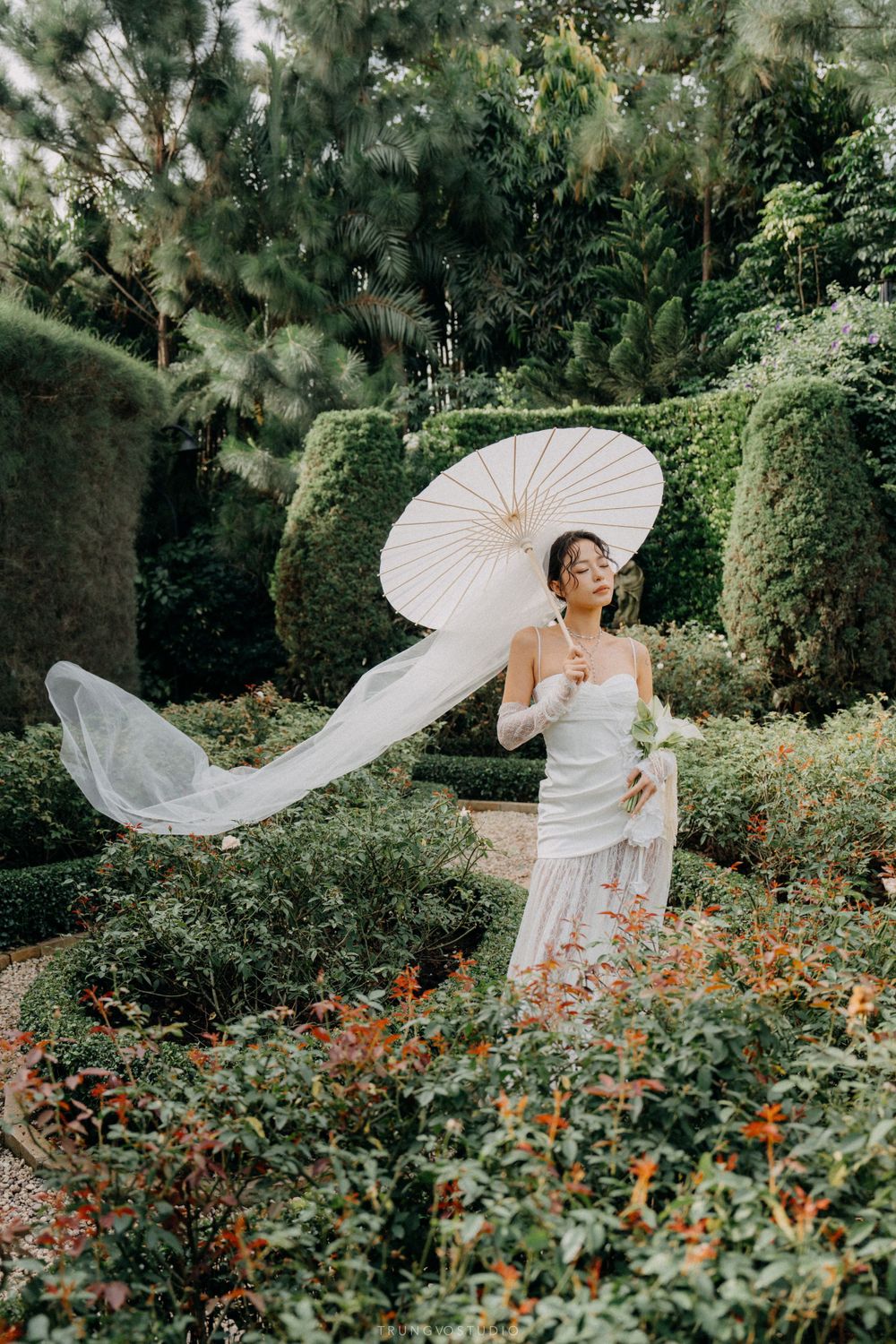 Bride in white gown and lace gloves poses with parasol as her veil flows in a lush garden landscape.
