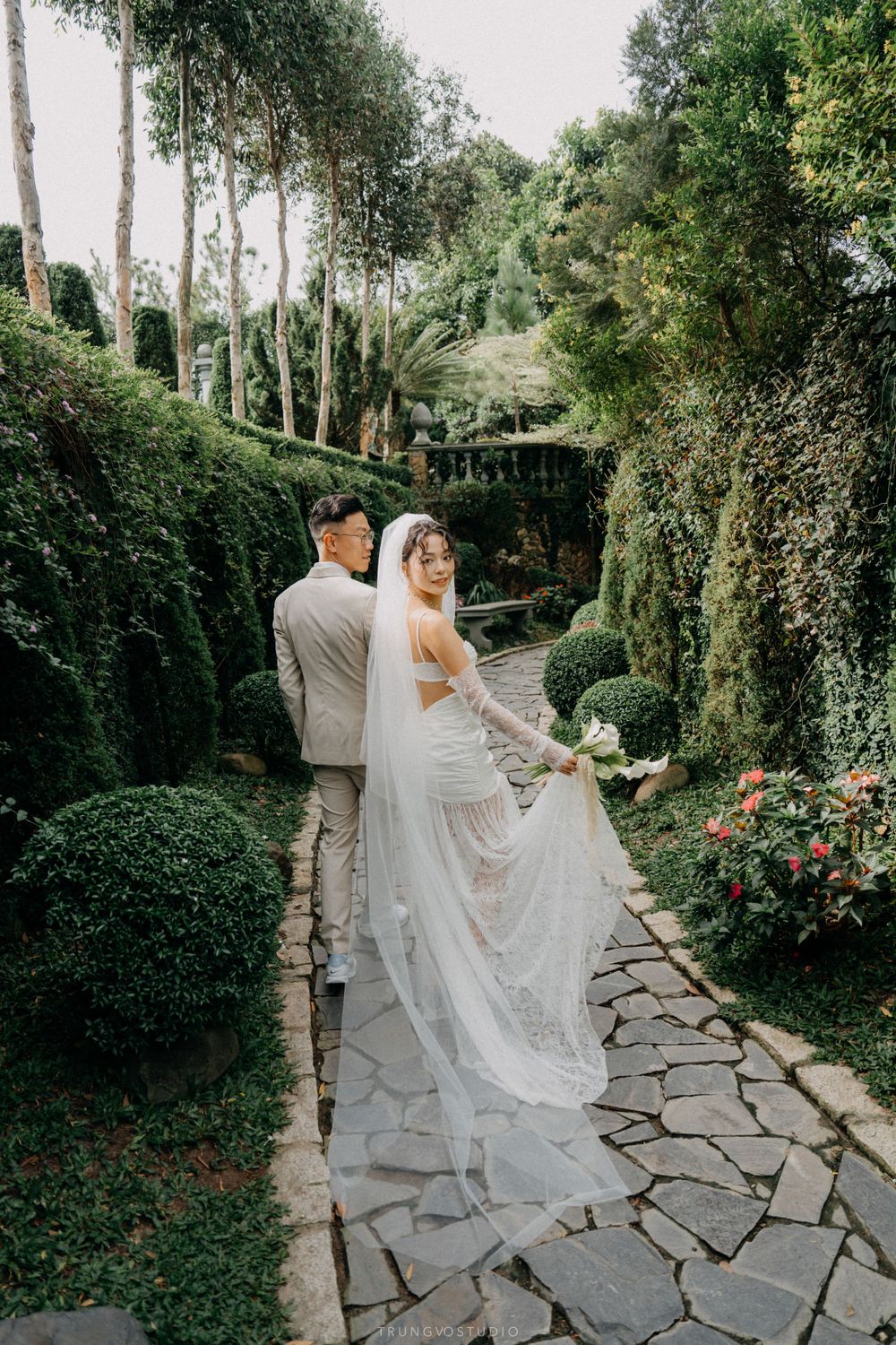 Bride and groom walk hand-in-hand along stone garden path surrounded by manicured hedges and colorful flowers.
