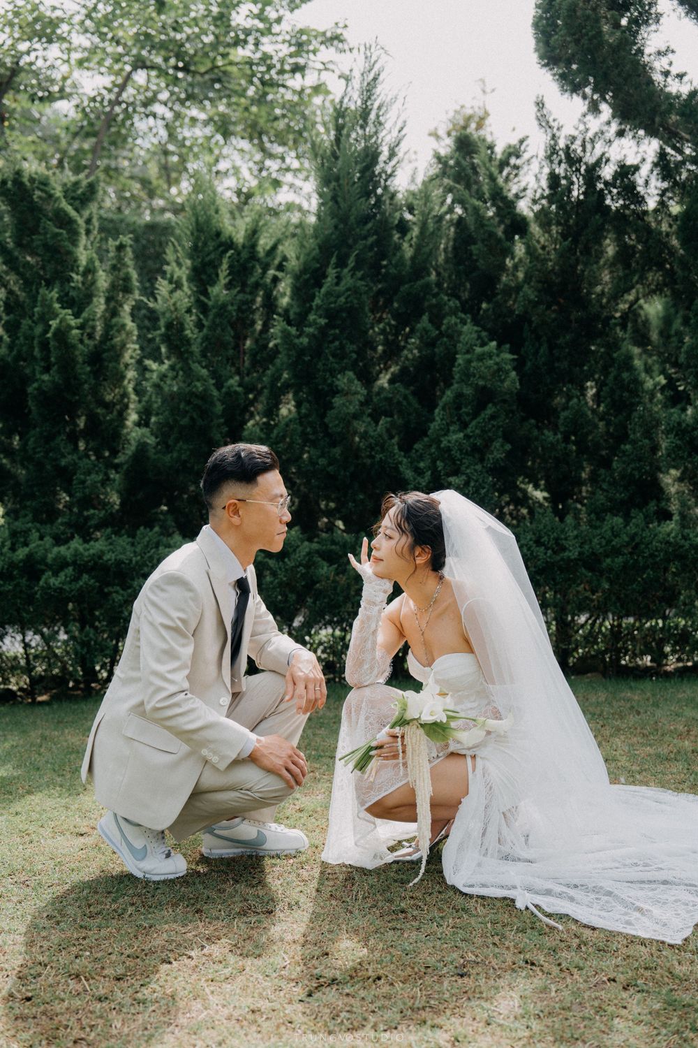 Bride and groom crouch facing each other on green lawn with tall evergreen trees in the background.
