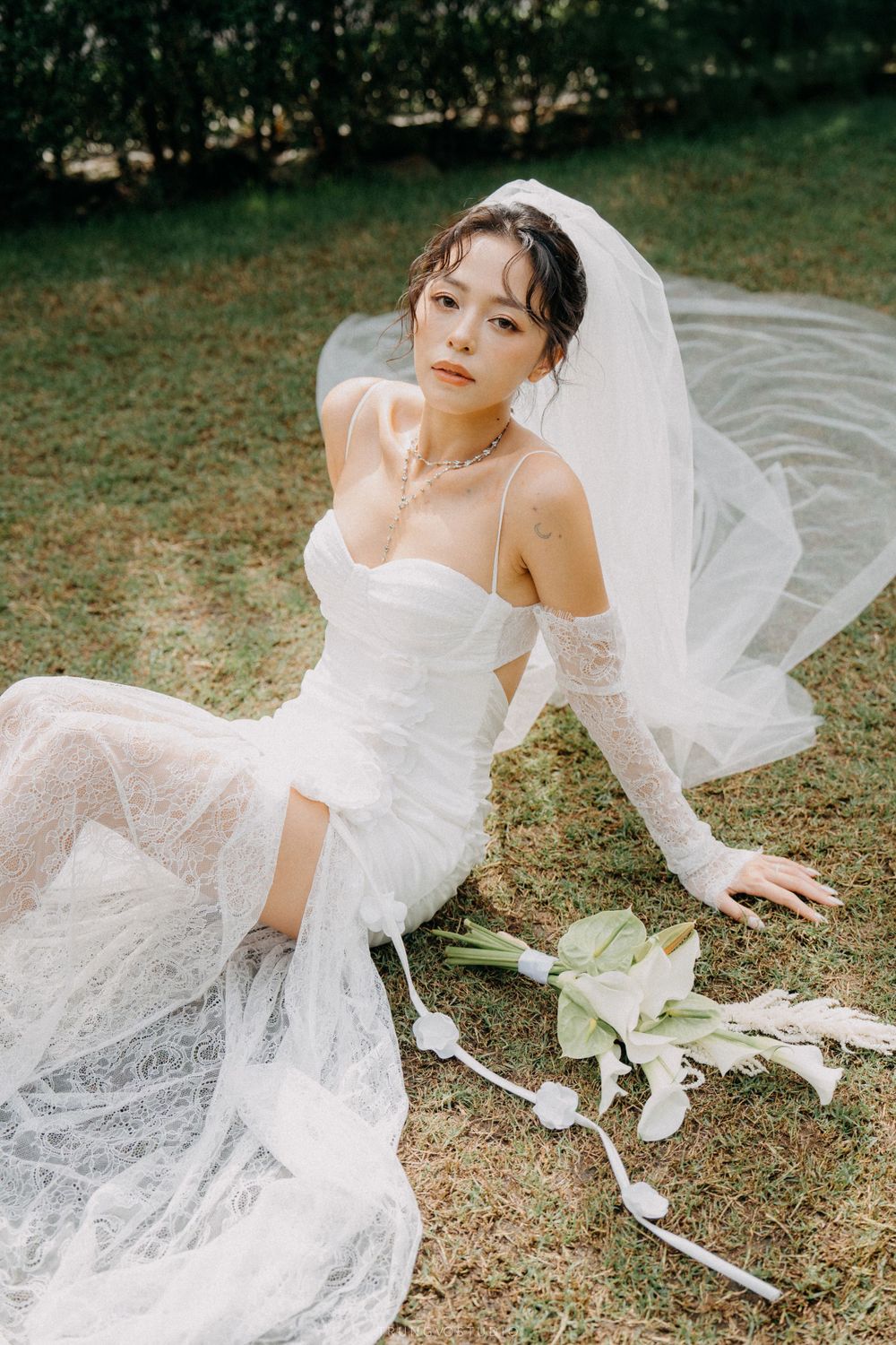 Elegant bride in white lace wedding dress and veil reclines on grass holding calla lily bouquet outdoors.