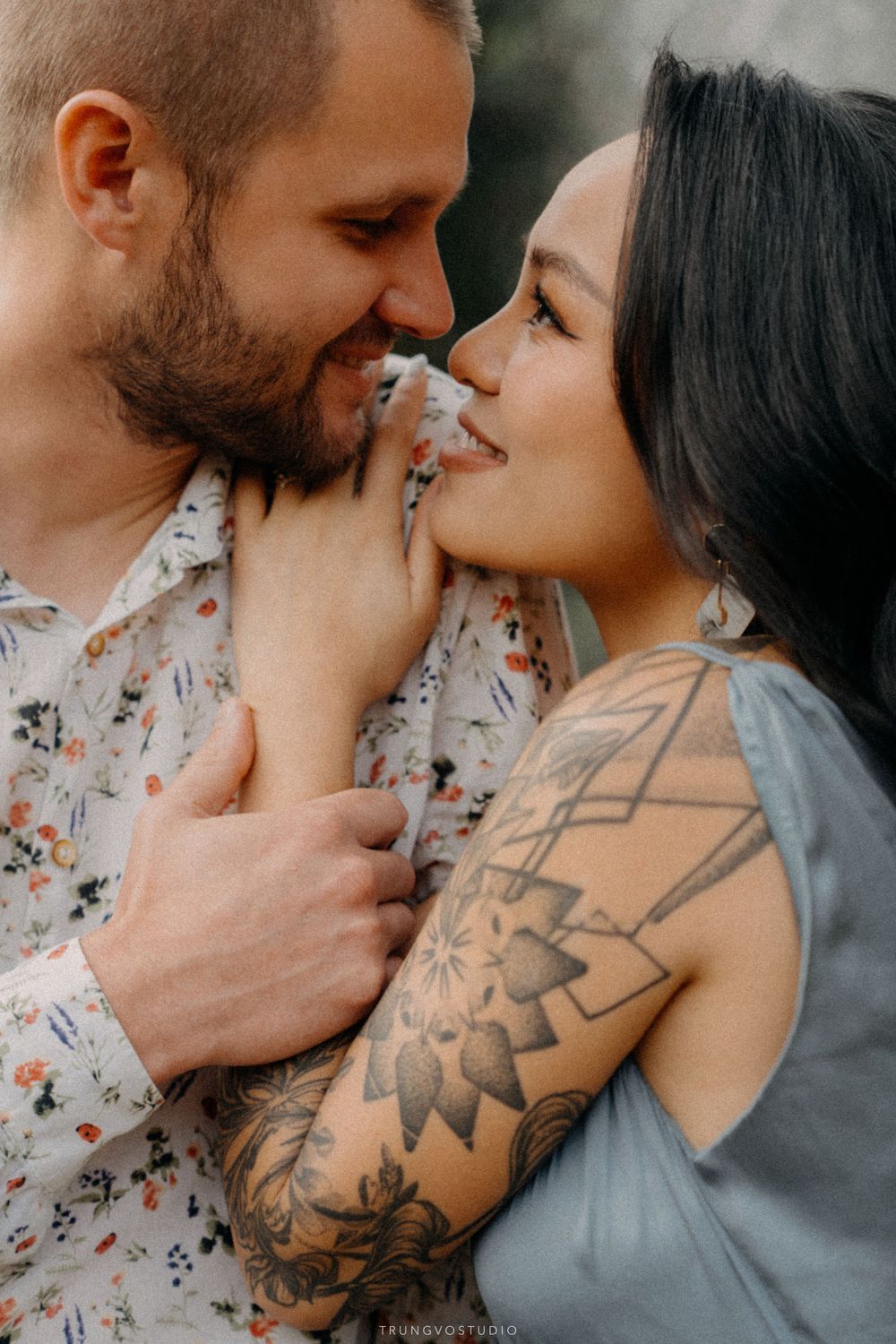 Tattooed couple in romantic close embrace, smiling at each other in soft natural light.