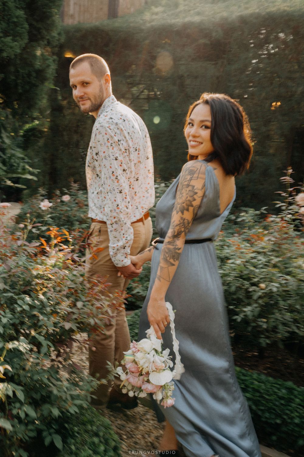 Smiling tattooed woman in gray dress holding bouquet turns back with man in floral shirt in garden.