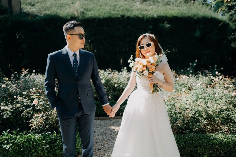 Bride and groom in sunglasses walk hand in hand through lush garden.