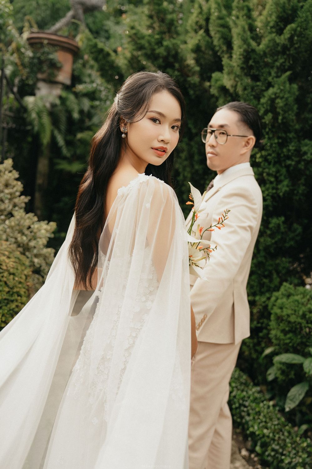 Elegant bride in white cape gown looks back while groom in beige suit stands among lush garden greenery.