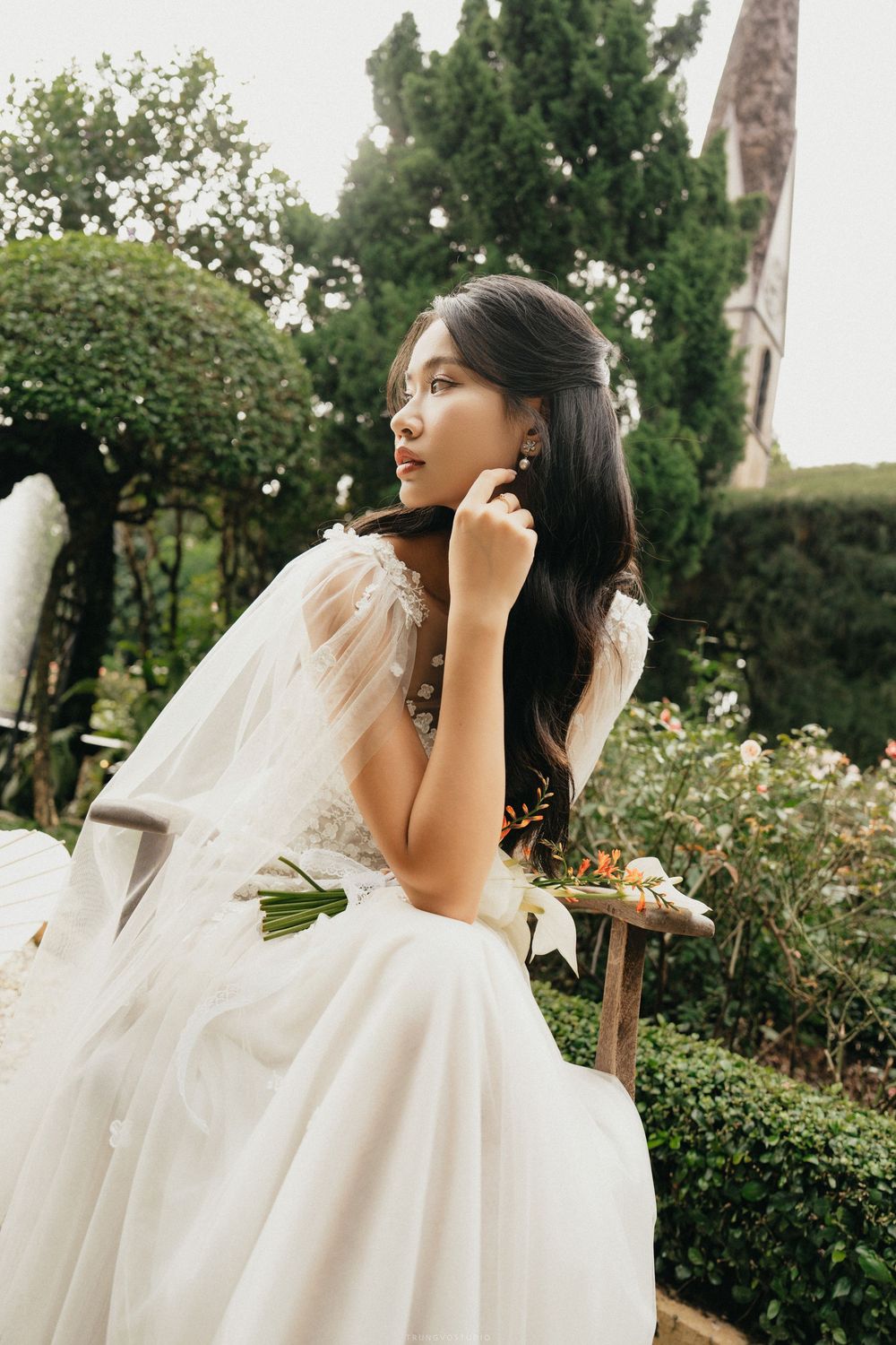 Elegant bride in tulle cape gown sits on bench holding bouquet in garden.