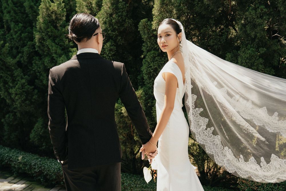 Bride in white gown with flowing lace veil holding groom's hand while looking back in garden.