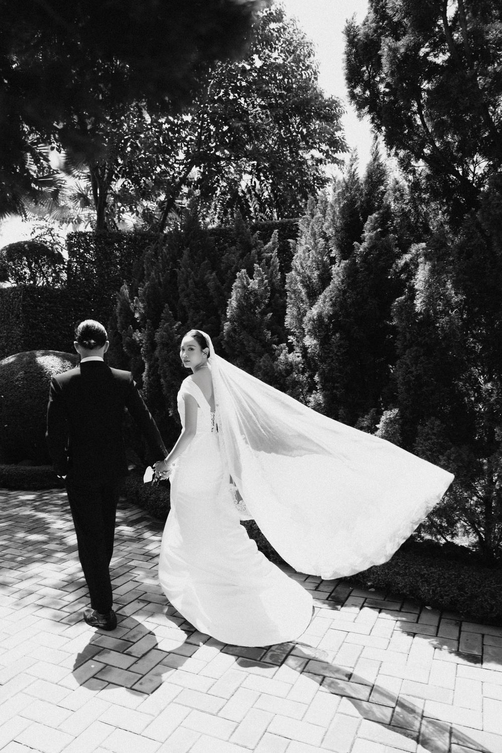 Black and white photo of bride and groom walking with veil billowing in the breeze.