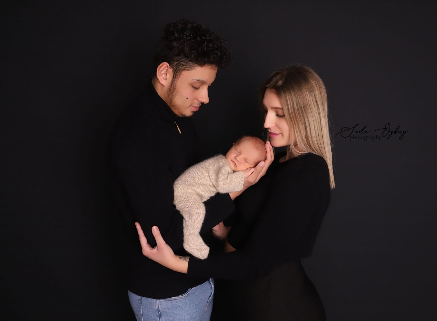 A tender family moment captured in a dark studio setting with a sleeping newborn baby.