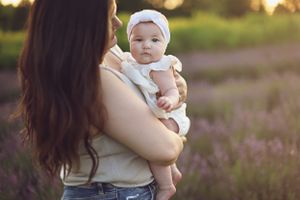 A woman holding a baby in white outfit against sunset in lavender field.