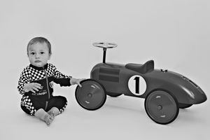 Baby boy in checkered outfit sits beside a vintage toy race car numbered 1 in black and white photo.