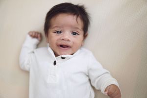 A baby wearing a white outfit smiles while lying on a light-colored background.