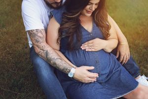 A couple sitting together on grass with hands resting on a baby bump, wearing denim and white attire.