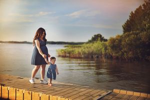 A parent and child stand together on a wooden dock at sunset overlooking a peaceful lake surrounded by greenery.