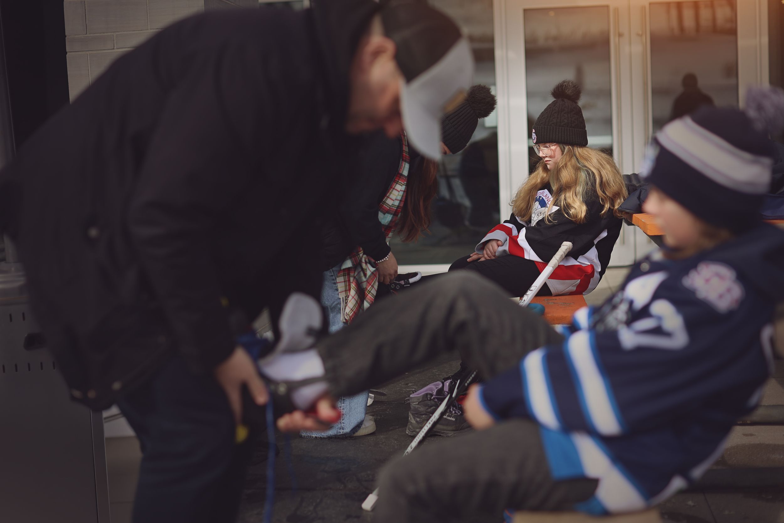 People sitting outdoors in winter clothing while a person assists with hockey skates on a cold day.