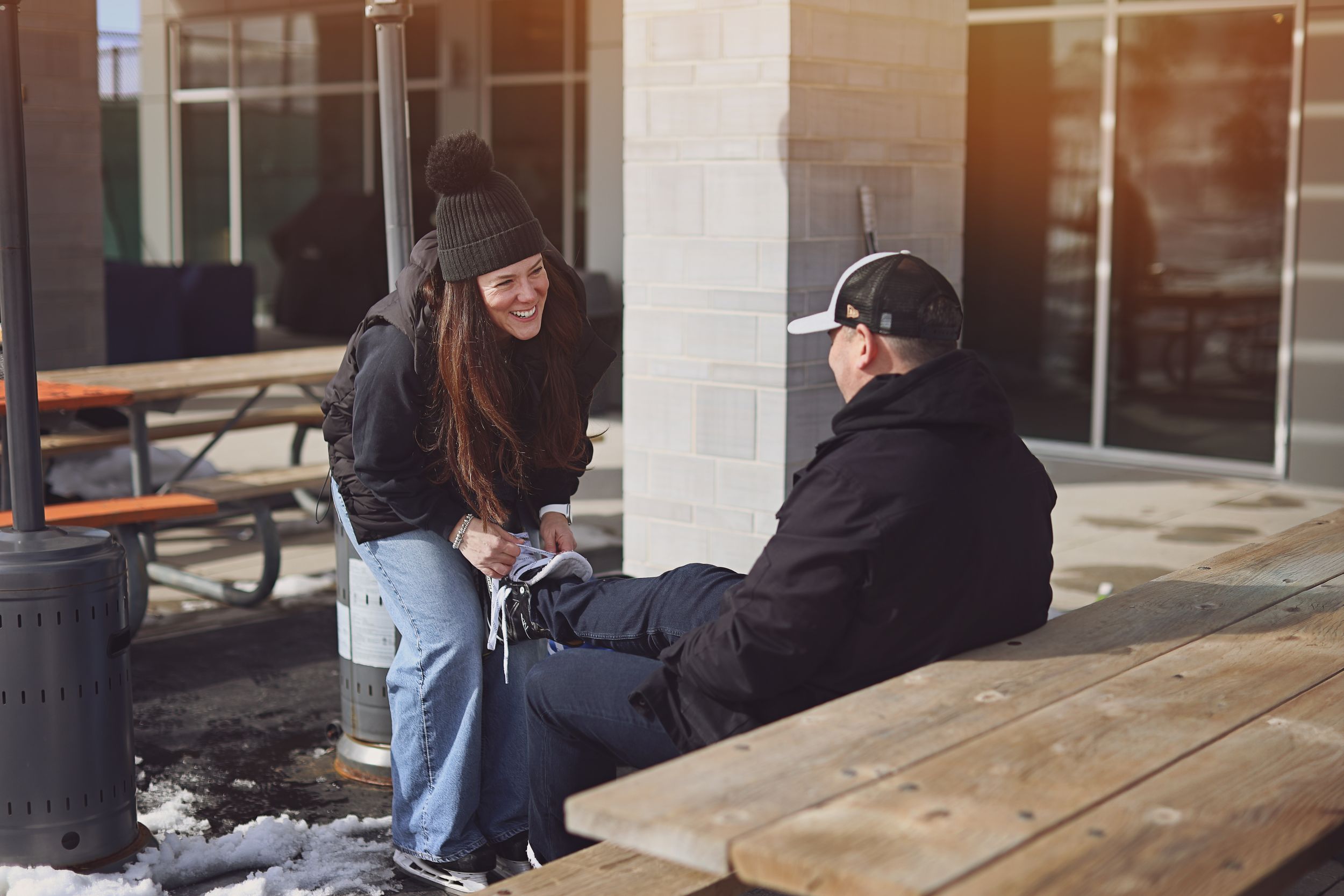 Two people in winter clothing chat and laugh at an outdoor wooden picnic table on a snowy day.