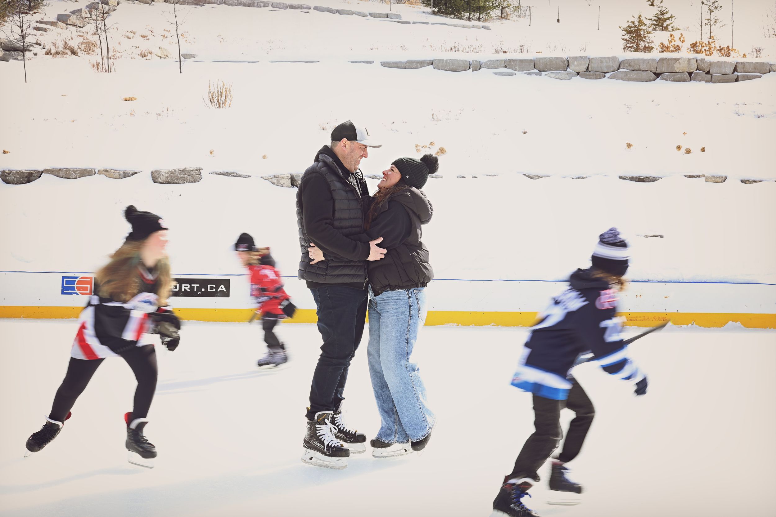 Couples and children ice skating on an outdoor rink surrounded by snow-covered landscape in winter.