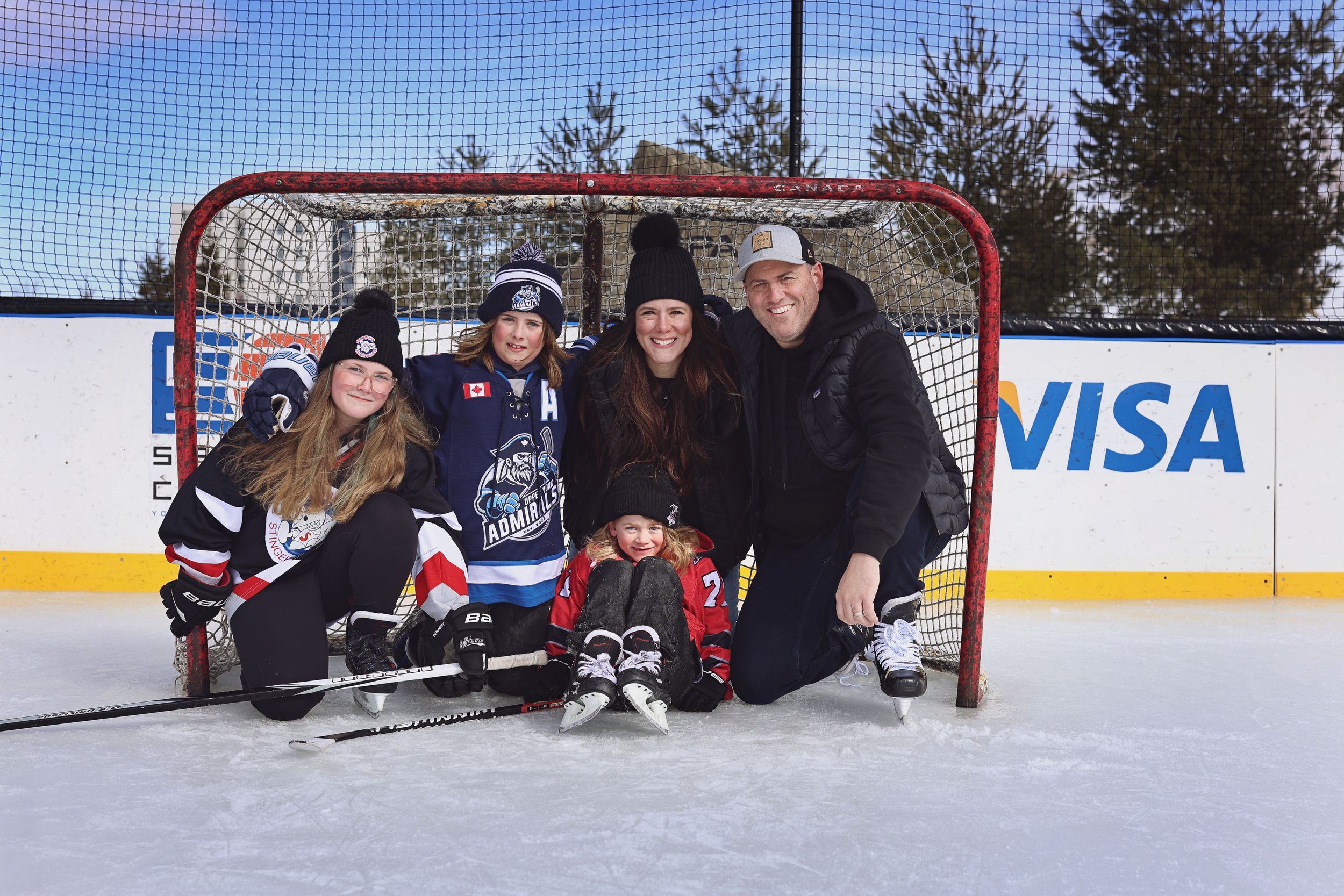 Family posing together on outdoor ice rink in front of hockey net, wearing jerseys and skates with Visa banner.