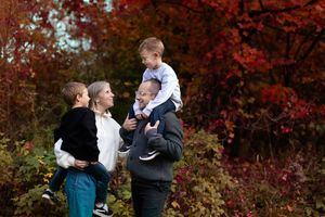 A family shares tender moments together in a park with vibrant red autumn foliage in the background.