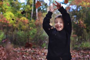 A joyful figure in black clothing plays with falling autumn leaves against a colorful fall forest backdrop.