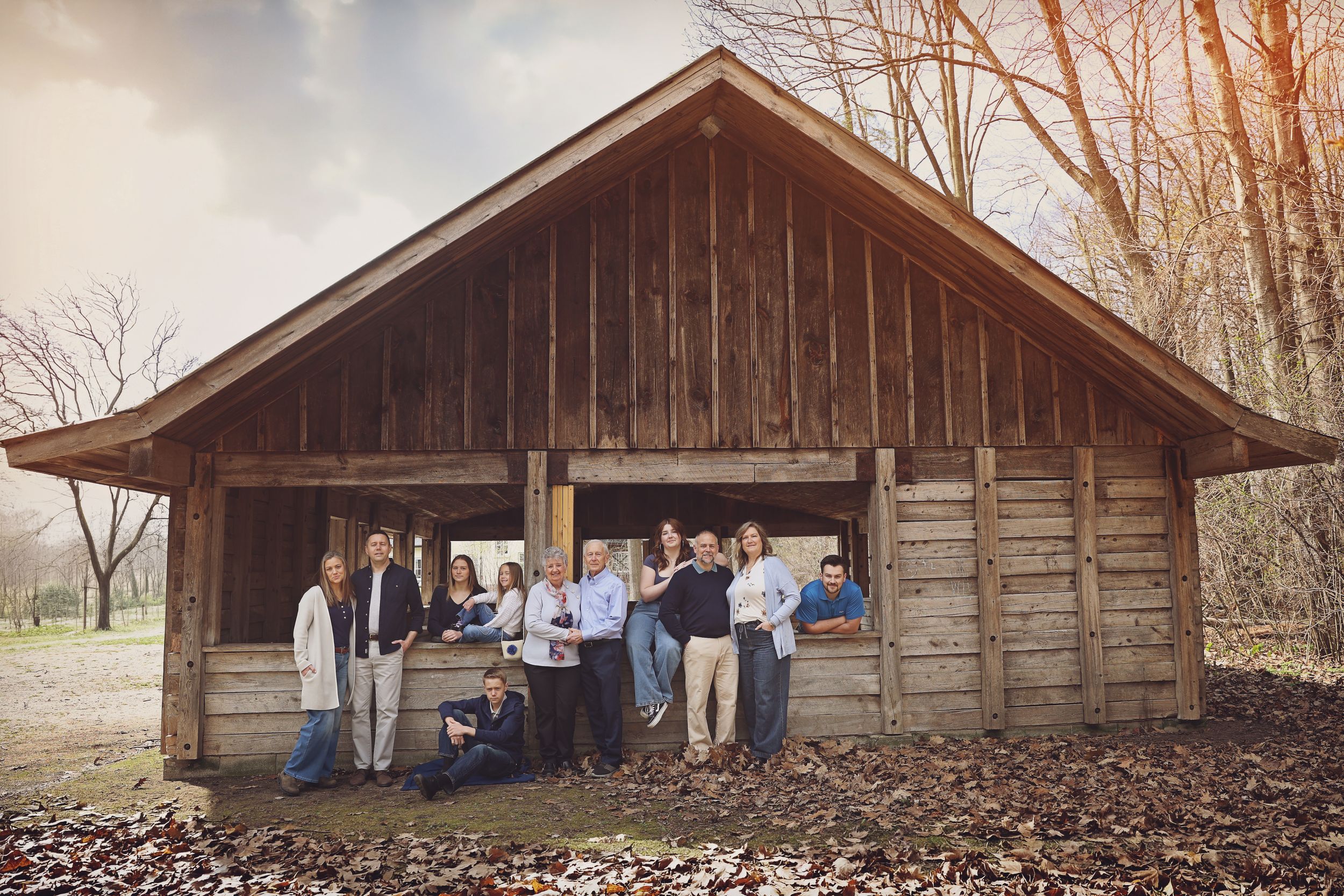 Large family group posing together inside a rustic wooden barn shelter surrounded by autumn trees.