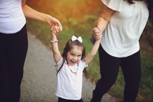 A joyful toddler in white holds hands with adults while walking outdoors.