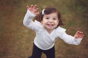A cheerful toddler in white raises arms with excitement while playing outdoors.