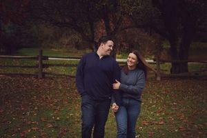 A couple laughs together while walking through a park on an autumn day with fallen leaves on the ground.