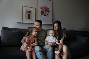 A family sits together on a dark grey couch in a living room with modern artwork on the wall.