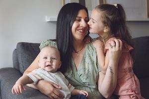 A family of three shares a tender moment together on a gray couch wearing sage green and pink clothing.