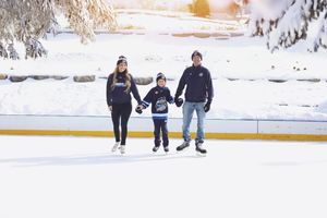 Family of three ice skating together on an outdoor rink surrounded by snowy trees in winter.
