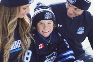 Young hockey fan smiles wearing team jersey and beanie, flanked by two adults in matching navy hockey apparel outdoors.