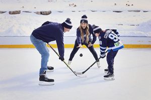 Three people playing outdoor ice hockey on a rink, two holding sticks at a face-off while a woman in a hat watches.