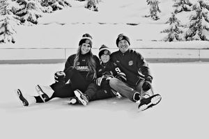 Family of three sitting on a snowy ice rink in winter gear, smiling together in a black and white photo.