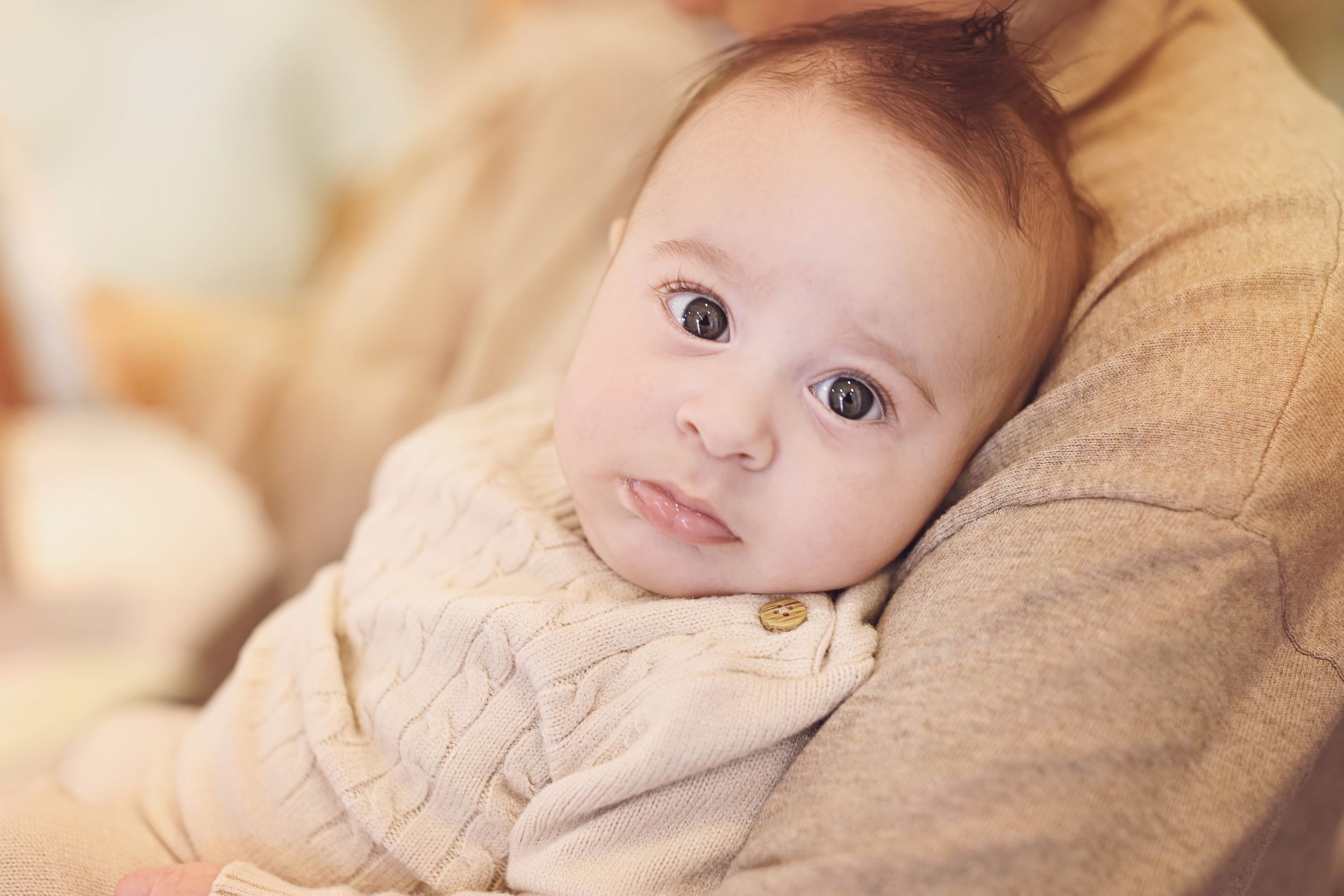 Adorable baby with big brown eyes resting on parent's shoulder in soft light.