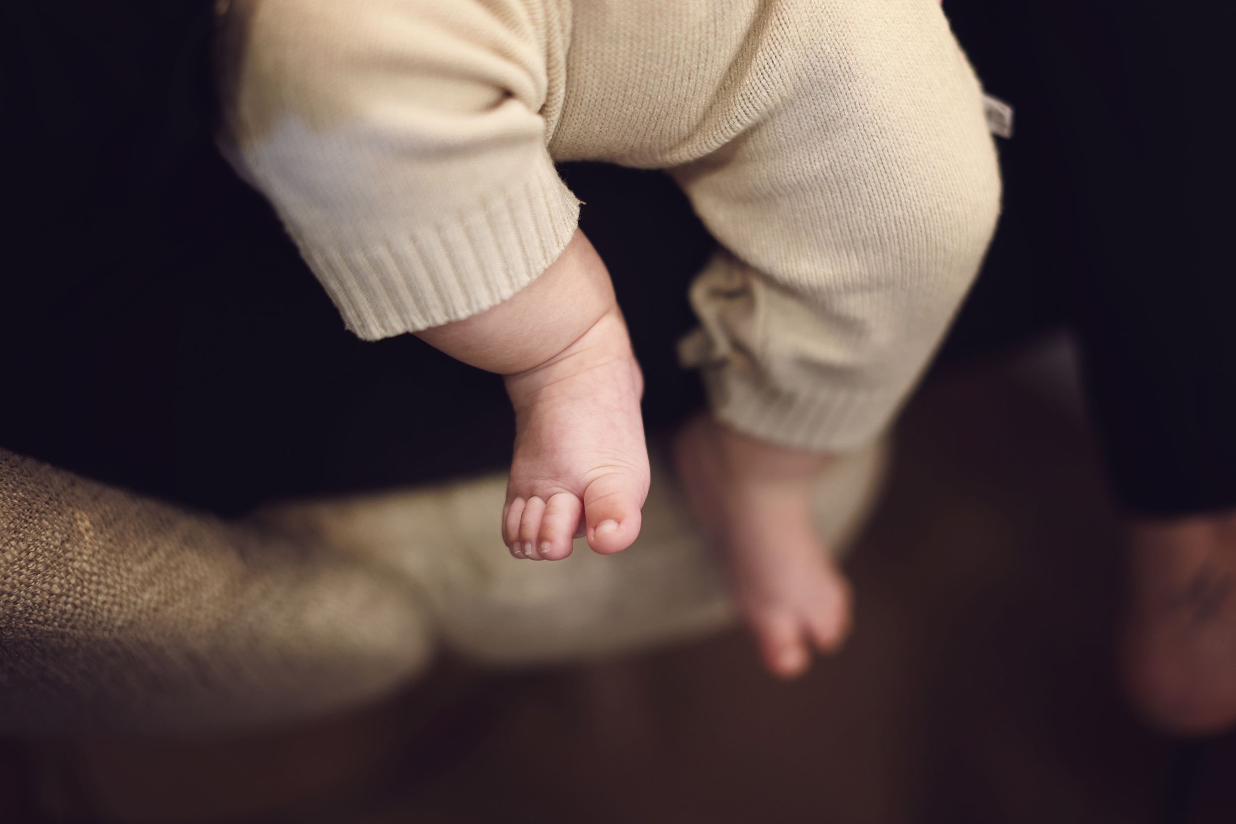 Close-up of a baby's tiny bare feet and chubby legs in cream knit pants, dangling in the air.