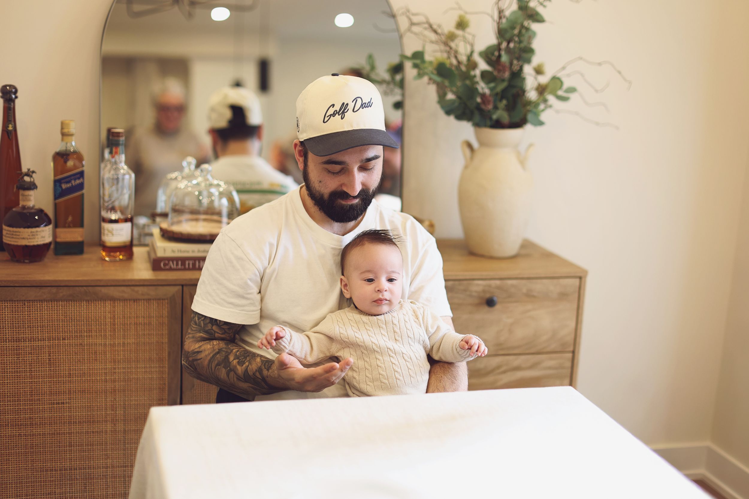 Tattooed man wearing a white cap tenderly holds a small baby in a cozy, warmly decorated room.